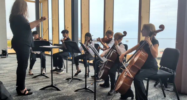A conductor leading young violinists and cellists in front of tall windows
